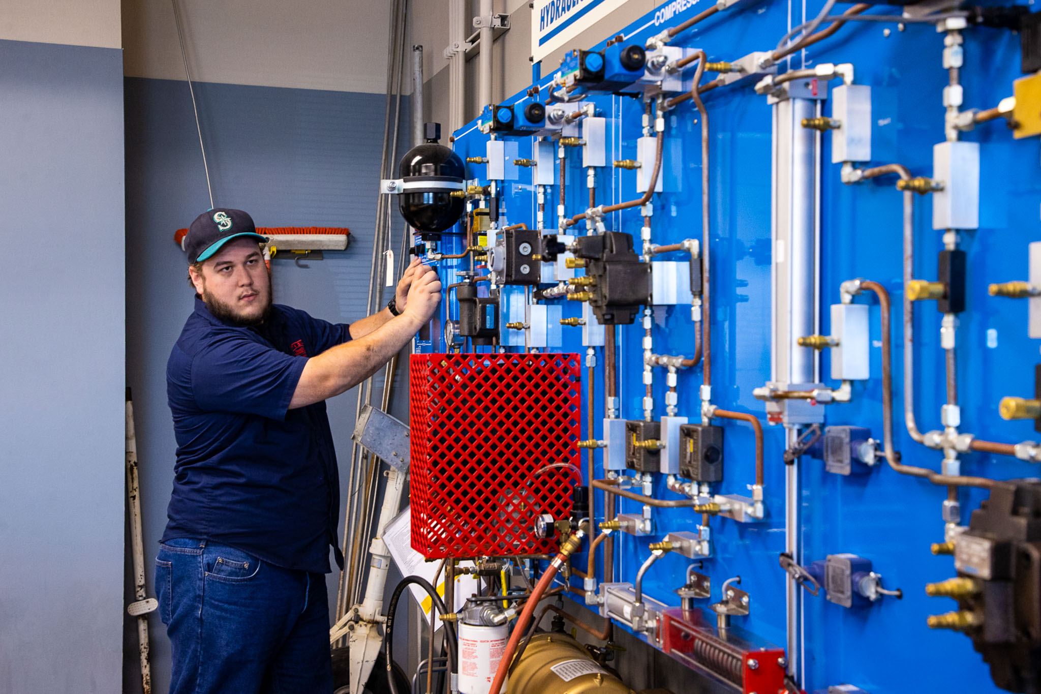 Student in a navy shirt and cap adjusts valves on a blue hydraulic training panel with gauges, pipes, and fittings in a technical lab setting.